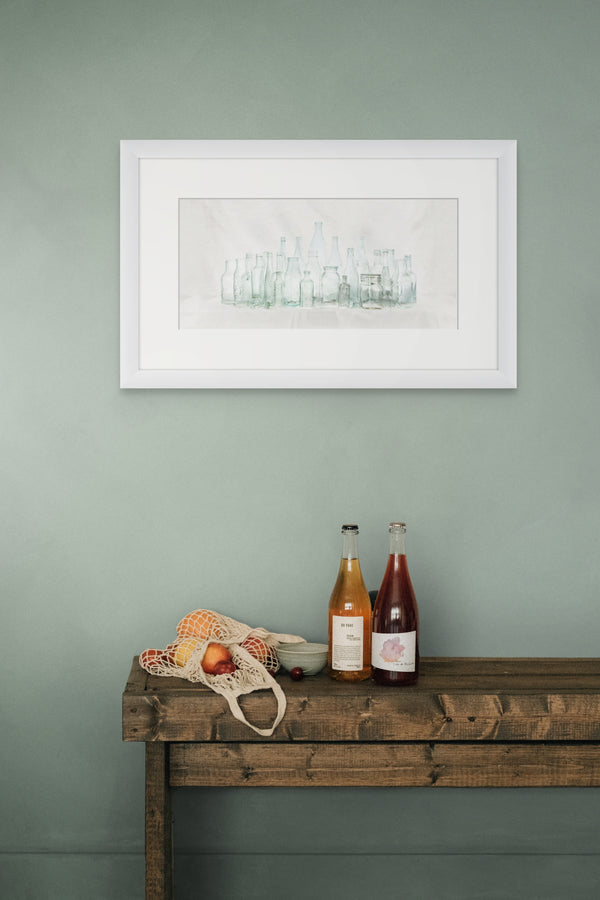 Framed artwork on a wall above a wooden table with bottles and fruits.